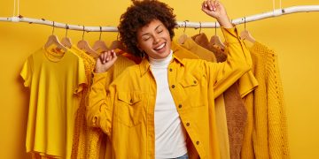 Carefree curly woman clothes buyer dances with happiness, raises arms, buys yellow clothing from new collection, rejoicing successful shopping day, being in high spirit, dances against outfits on rack