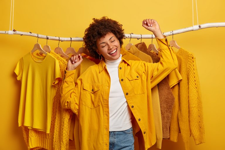 carefree curly woman clothes buyer dances with happiness raises arms buys yellow clothing from new collection rejoicing successful shopping day being high spirit dances against outfits rack