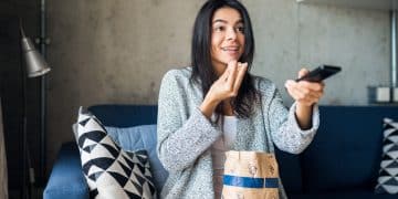 attractive smiling happy woman sitting on sofa at home watching tv movies online holding remote control eating popcorn wih emotional surprised face expression